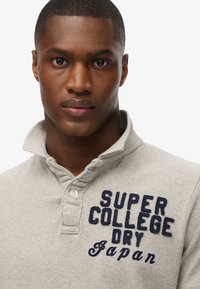 Grijze polo-shirt met korte mouwen en een kraag. Bevat marineblauwe geborduurde tekst: "SUPER COLLEGE DRY Japan." Zachte textuur.