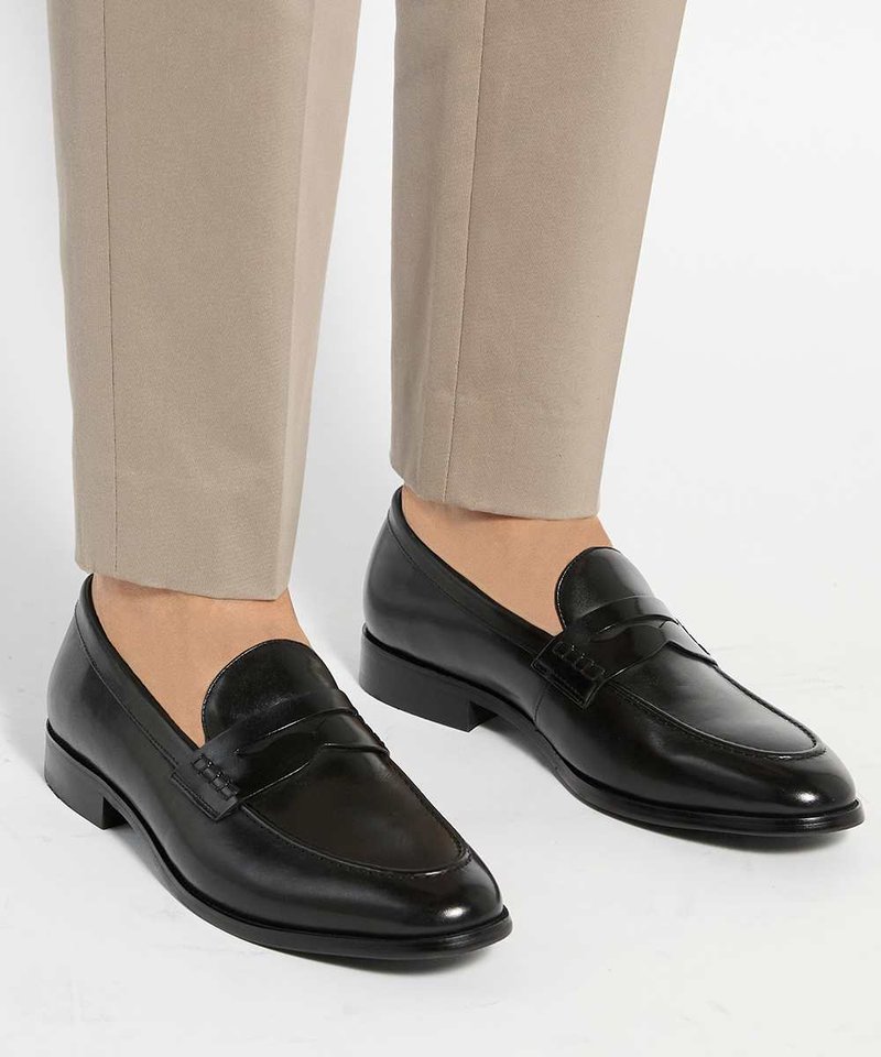 Mocassins en cuir noir sans lacets portés avec un pantalon beige, mettant en avant le design élégant et poli des chaussures ainsi que leurs talons bas.