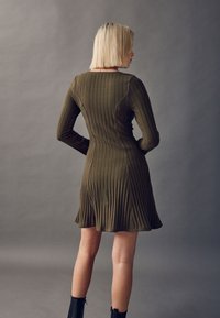 Langärmeliges, olivgrünes, geripptes Kleid mit einem taillierten Oberteil und ausgestelltem Rock. Glatte Textur mit vertikalen Falten. Getragen mit schwarzen Stiefeletten.