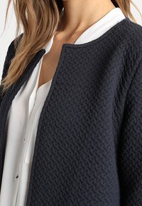 Navy strukturierte Jacke mit Rundhalsausschnitt, offenem Design und weichem Stoff. Getragen über einem weißen Hemd mit Knöpfen.
