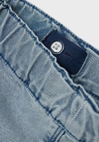 Los pantalones de mezclilla azul claro tienen una cintura deshilachada y un parche textil azul marino con un botón transparente, mostrando sutiles detalles de costura.