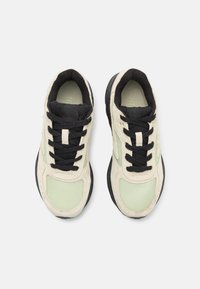Baskets de sport avec une tige crème, des lacets noirs, des détails en maille et une semelle en caoutchouc noir texturé. Accents verts sur les côtés.