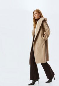 Abrigo de lana beige con una gran capucha y botones de doble, combinado con pantalones oscuros de campana y tacones negros de punta.