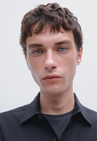Ein junger erwachsener Mann mit strukturiertem dunklem Haar und blauen Augen trägt ein schwarzes Hemd mit Kragen und ein dunkles Inneres Hemd, minimaler Hintergrund.