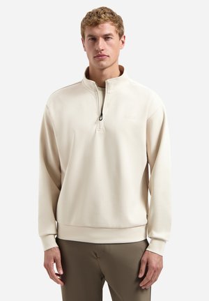 Jeune homme portant un pull beige à fermeture éclair quart de zip et un pantalon vert olive, debout devant un fond blanc uni.