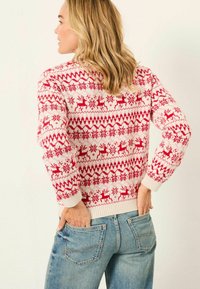 Frau mit blonden Haaren, die seitlich steht und einen weißen und roten festlichen Pullover mit Rentier- und Schneeflockenmuster sowie eine blaue Jeans trägt.