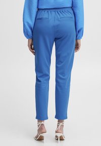 Pantalons bleus à taille élastique, jambes ajustées, tissu lisse, poches arrière, associés à des sandales à talons hauts blanches avec des liens autour de la cheville.