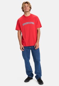 Camiseta de algodón roja con texto gráfico azul "Quiksilver", de manga corta y cuello redondo, combinada con jeans de mezclilla azules y zapatos negros de tipo slip-on.