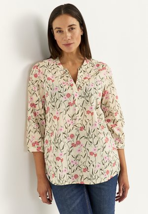 Vrouw die tegen een muur staat, gekleed in een crèmekleurige blouse met roze bloemenprint en driekwartmouwen, gecombineerd met een blauwe spijkerbroek.