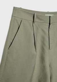 Pantalon en lin vert olive clair avec plis devant, poches latérales, passants de ceinture et fermeture éclair dissimulée.