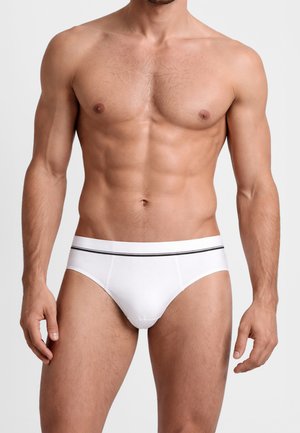 Torso maschile muscoloso indossando slip bianchi con una fascia in vita a righe nere, in piedi contro uno sfondo chiaro e semplice.