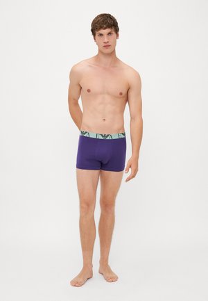 Lila Boxer-Shorts aus glattem Stoff, mit einem kontrastierenden mintgrünen Bund, der ein Logomuster zeigt. Das Modell steht barfuß.