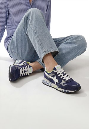 Person wearing blue sneakers with white accents and light blue jeans, sitting cross-legged on a white floor.