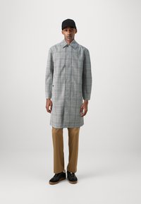 Cappotto lungo a scacchi grigi con colletto ampio, chiusura a bottoni e maniche lunghe, abbinato a pantaloni beige e sneakers nere.