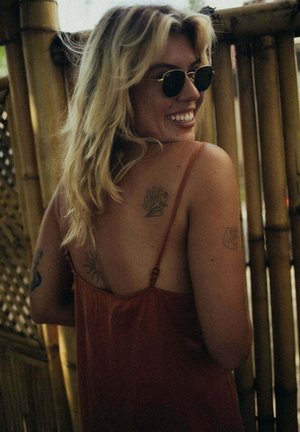 Letnia sukienka w kolorze rdzy z cienkimi ramiączkami; gładki materiał; prezentowana na tle bambusa; widoczne tatuaże na ramieniu i plecach.