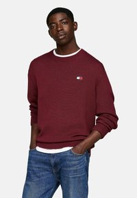 Suéter de punto burdeos con cuello redondo y puños acanalados; pequeño parche con logo en el pecho; usado sobre una camisa blanca; combinado con jeans azules.