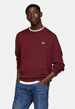 Suéter de punto burdeos con cuello redondo y puños acanalados; pequeño parche con logo en el pecho; usado sobre una camisa blanca; combinado con jeans azules.