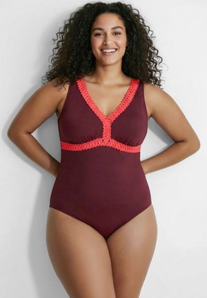 Smilende kvinde med krøllet hår iført en ærmeløs vinrød badedragt med klar rød kant, stående med hænderne bag ryggen.