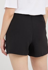 Czarne szorty z gładkiego materiału, w luźnym kroju i lekko zaokrąglonym dole. Widok z tyłu, bez widocznych elementów metalowych.