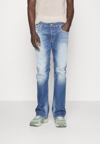 Homme portant un jean bleu clair délavé, un haut beige en tricot et des baskets vertes et blanches, debout avec une main dans la poche.