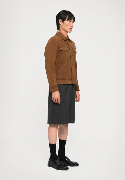 Brązowa zamszowa kurtka z kołnierzem, przednimi kieszeniami i dopasowanym krojem. Połączona z szarymi szortami oraz czarnymi mokasynami i skarpetkami.