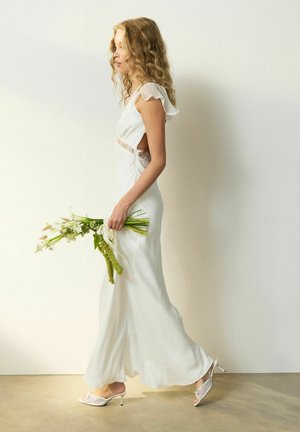 Sieviete baltā bezroku kleitā ar mežģīņu detaļām tur ziedu pušķi, staigājot pret vienkāršu gaišu fonu.