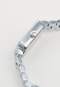 Silberner Edelstahl-Uhr mit rechteckigem Gehäuse, strukturiertem Metallarmband und strukturiertem Kranz an der Seite zur Einstellung.