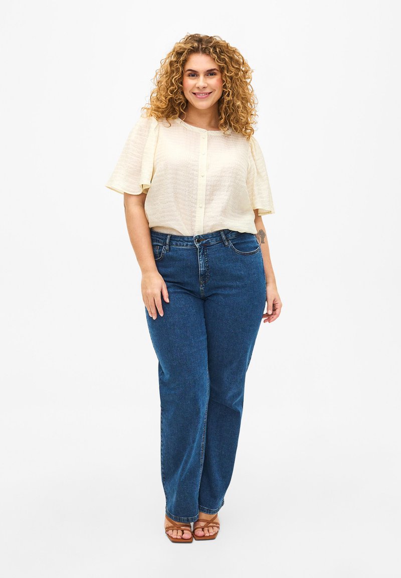Blusa di crema chiaro a maniche corte con un motivo testurizzato e bottoni frontali, abbinata a jeans blu a gamba dritta e sandali marroni con cinturini.