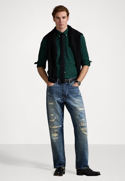 Svart quilted vest, grønn rutete skjorte, slitt blå jeans med lysere lapper, svarte sko og et klokke, stående i en nøytral setting.