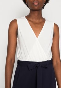 Top branco sem mangas com um design em V, combinado com calças azul-marinho de cintura alta com um laço cinto na cintura e bolsos laterais.