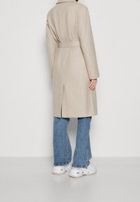 Béžový kabát z vlněné směsi s páskem v pase, dlouhými rukávy a rozparkem na zadní straně, kombinovaný se světle modrými zvonovými džíny a bílými teniskami.