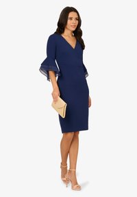 Robe ajustée bleu marine avec un design enroulé, manches cloche transparentes et mi-longue. Associée à une pochette beige et des sandales à talons nudes.