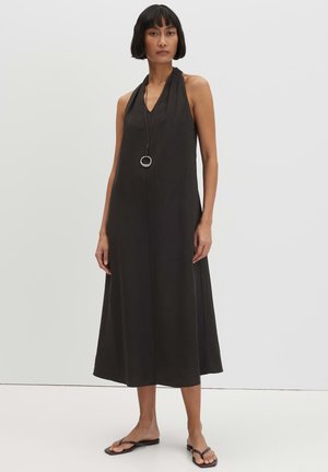 Vrouw met kort donker haar die een mouwloze zwarte halter midi-jurk draagt, een zwarte hangerketting en zwarte platte sandalen, staand tegen een witte muur.