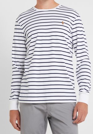 Homme portant une chemise blanche à manches longues avec des rayures horizontales bleu marine et un petit logo brodé de cheval sur la poitrine, associée à un pantalon gris clair.