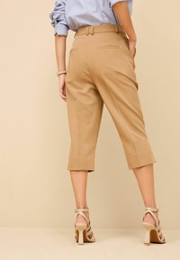 Hellbraune, tapered Hosen mit cropped Saum und Gesäßtaschen, kombiniert mit beigen, geschnürten High-Heels. Glatte Stofftextur.