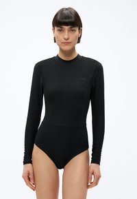 Kobieta o krótkich ciemnych włosach, ubrana w dopasowany czarny prążkowany body z długim rękawem na jednolitym białym tle.