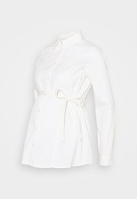 Vit amningsskjorta med långa ärmar, knäppning fram och knytning vid midjan, tillverkad av ett mjukt och lätt tyg.