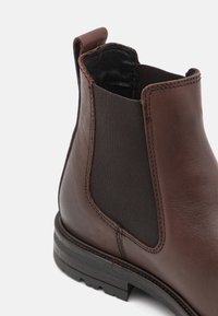 Botín Chelsea de cuero marrón con panel elástico en los lados, tira en el talón y suela texturizada negra sobre un fondo blanco.