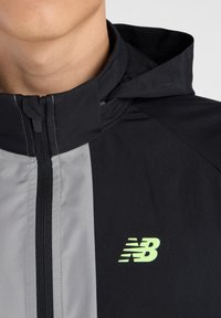 Zwarte en grijze windbreaker jas gemaakt van lichtgewicht materiaal, met een rits aan de voorkant, een capuchon en een neon groene logo op de borst.