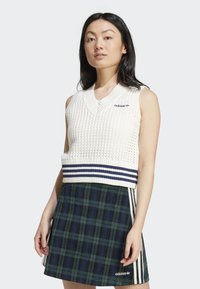Mulher vestindo um colete de malha branco sem mangas com riscas em azul-marinho e um logo da Adidas, combinado com uma saia xadrez verde e azul-marinho com riscas da Adidas.