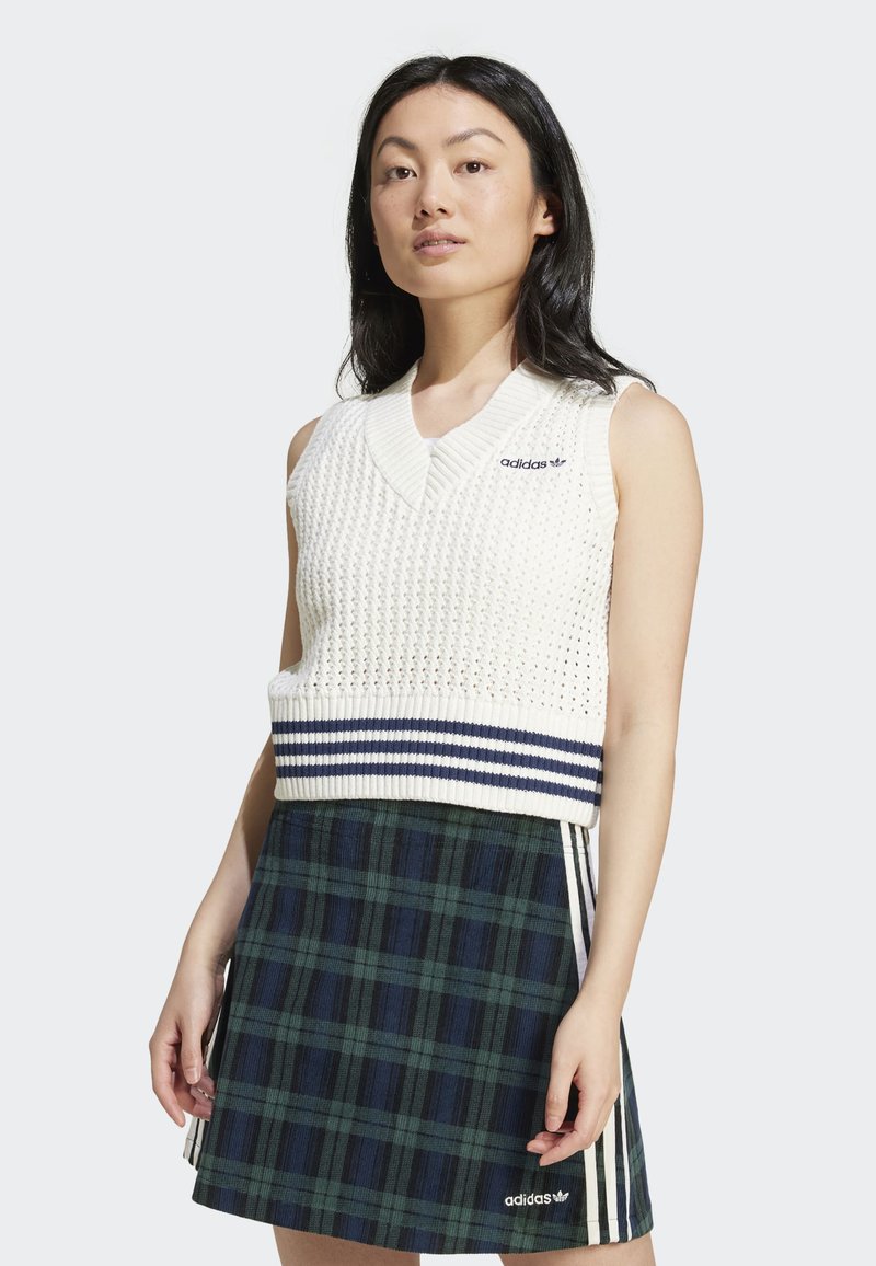 Mulher vestindo um colete de malha branco sem mangas com riscas em azul-marinho e um logo da Adidas, combinado com uma saia xadrez verde e azul-marinho com riscas da Adidas.