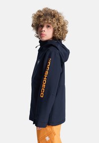 Granatowa kurtka z kapturem z zamkiem i pomarańczowym logo na rękawie, w zestawie z pomarańczowymi spodniami. Posiada opływowy kształt i miękką teksturę.