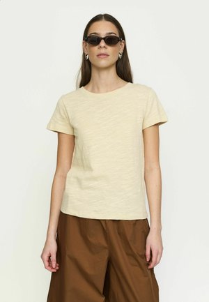 Žena v béžovom tričku s krátkym rukávom, hnedých širokých nohaviciach, tmavých oválnych slnečných okuliaroch a strieborných kruhových náušniciach, stojaca pred jednoduchým pozadím.