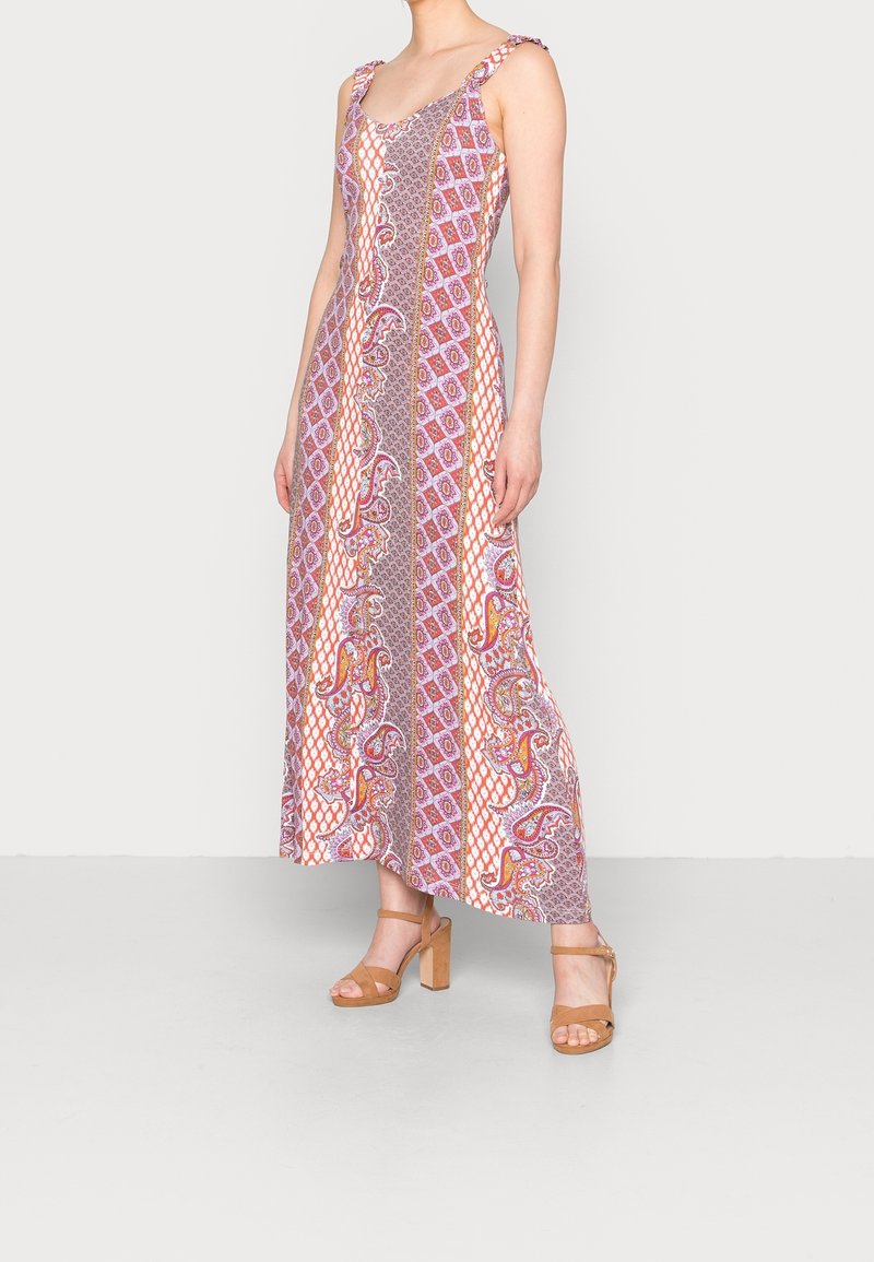 Femme portant une robe longue sans manches multicolore avec des rayures verticales à motifs et des sandales marron à talons hauts sur un fond uni.