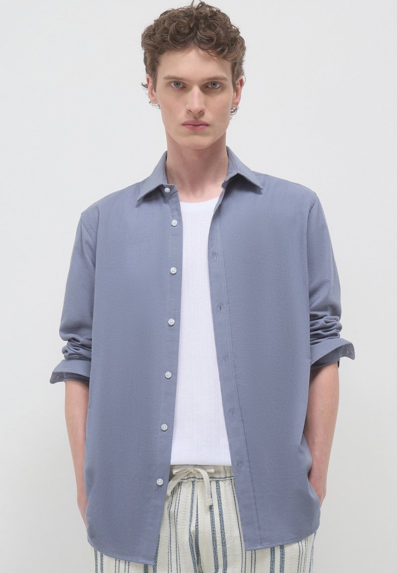 Junger Mann mit lockigem Haar, der ein hellblaues, kurzärmliges Hemd über einem weißen Oberteil und gestreiften Bundhose trägt, vor einem neutralen Hintergrund.