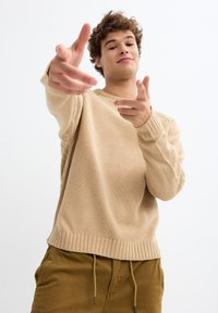 Suéter de punto beige con cuello redondo, puños acanalados y dobladillo. El modelo posa con los dedos señalando, vistiendo pantalones caqui claros.