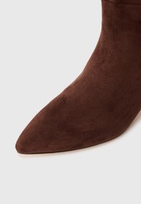 Barna velúr, hegyes orrú bokacsizma sima textúrával és zökkenőmentes kialakítással, magas sarkú, látható hardver vagy mintázat nélkül.