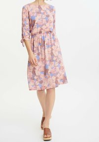 Kobieta ubrana w różowo-lawendową sukienkę w kwiaty do kolan, z rękawami 3/4 i brązowymi sandałami na koturnie, stojąca na białym tle.