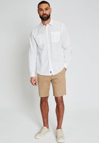 Camisa blanca de manga larga con botones y dos bolsillos en el pecho, combinada con pantalones cortos caquis y zapatillas blancas. Tejido suave, ajuste relajado.