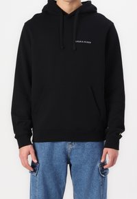 Homme portant un hoodie noir Lyle & Scott avec poche avant et un jean cargo bleu, se tenant contre un fond blanc uni.
